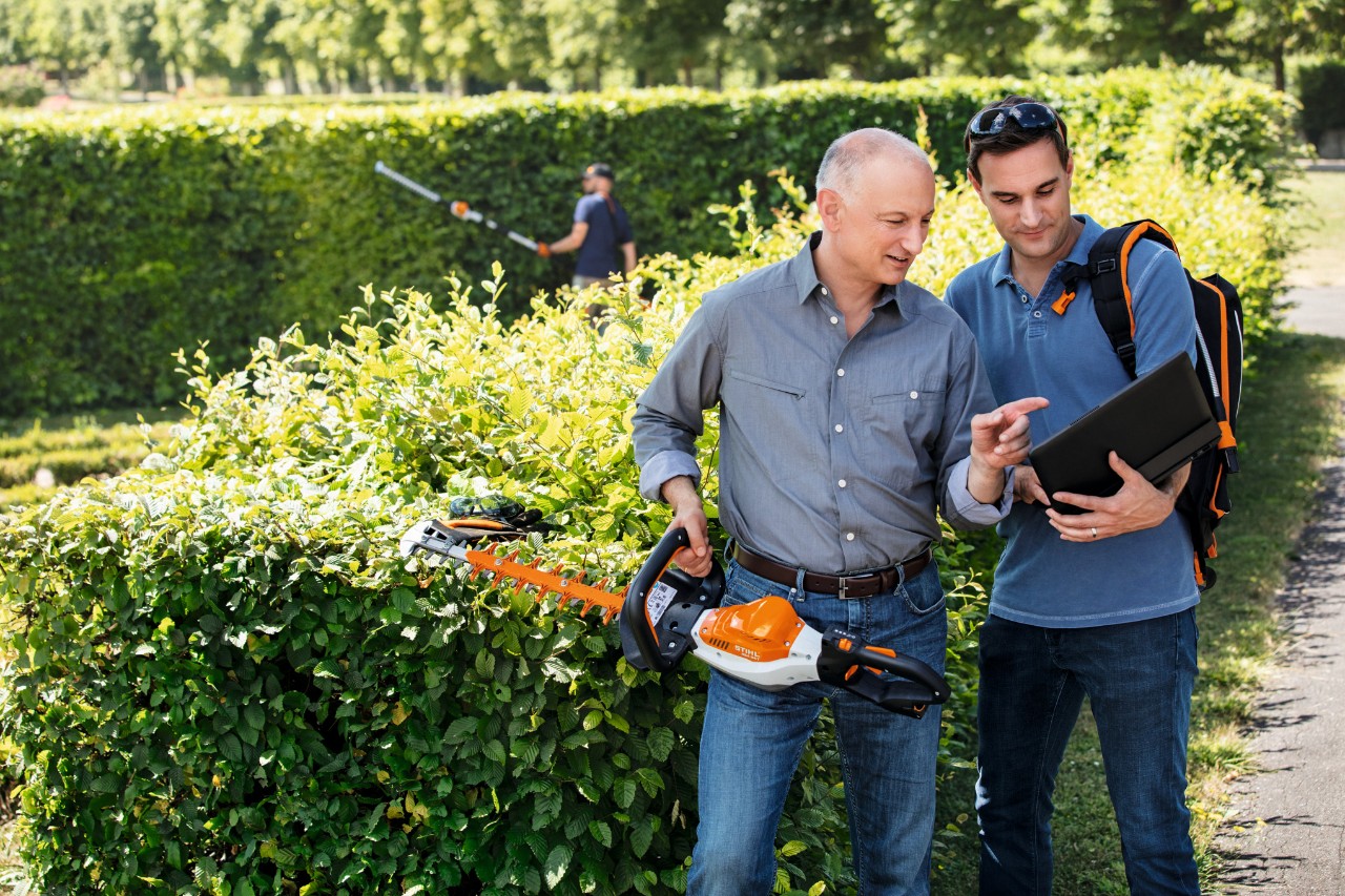 Д-р Николас Щил държи ножица за жив плет STIHL в ръката си и разговаря с човек, който стои до него и държи лаптоп.