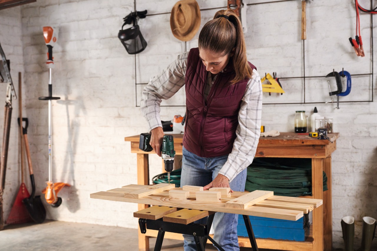 Жена с карирана риза и коса, вързана на конска опашка, завинтва летви към подпорни опори на работна маса, на заден план – стена с оборудване