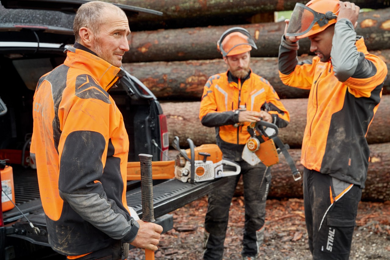 Трима мъже в защитно облекло STIHL пред дървени купчини се подготвят за работа с верижен трион STIHL