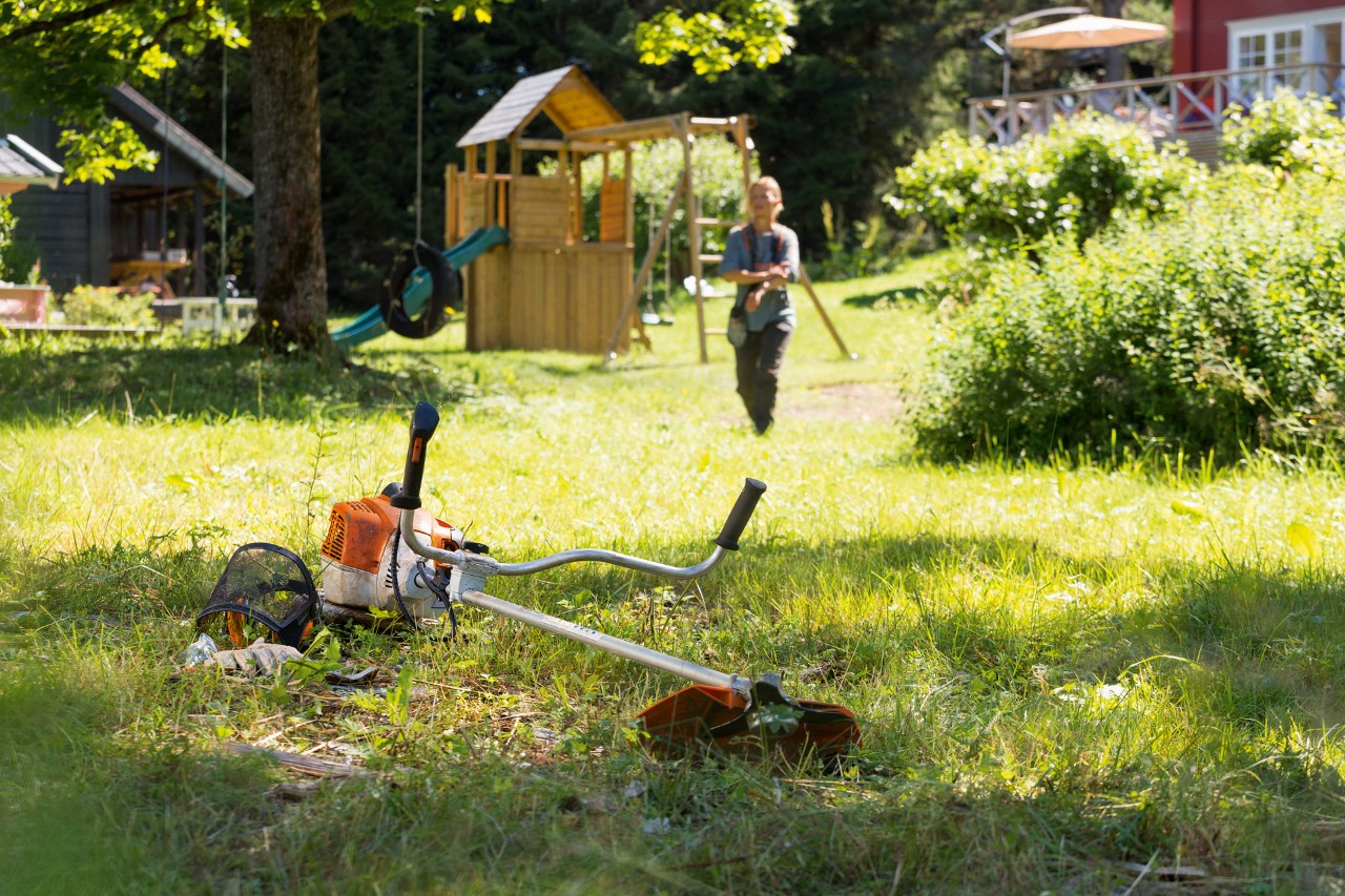Моторна коса STIHL FS 240 лежи в тревата, на заден план мъж се подготвя за техническо обслужване