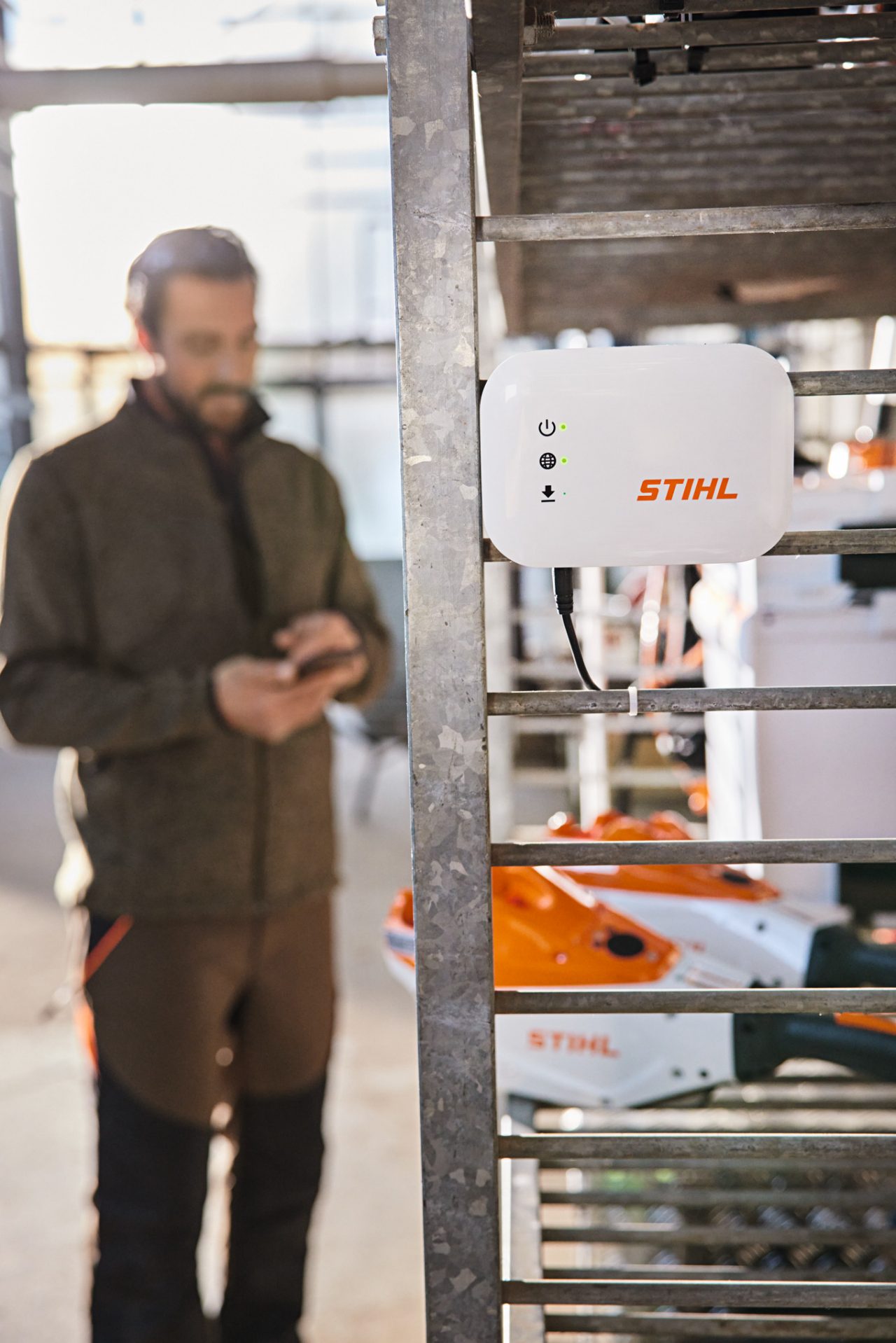 Кутия STIHL Connected виси на сервизен регал. На заден план се вижда градински и ландшафтен специалист, който работи със своя смартфон. 
