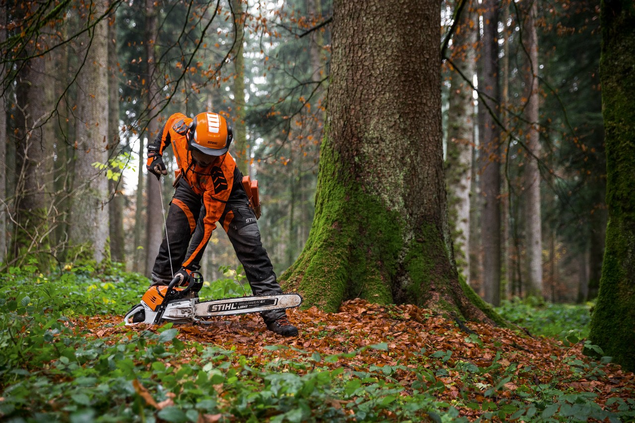 Мъж в предпазно облекло стартира бензинов верижен трион STIHL MS 500i в разлистена гора на земята