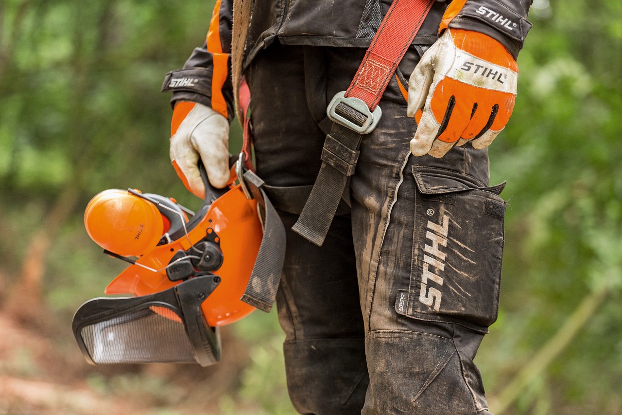 Човек с панталон за защита срещу срязване STIHL и ръкавици STIHL държи предпазна каска STIHL в ръка 