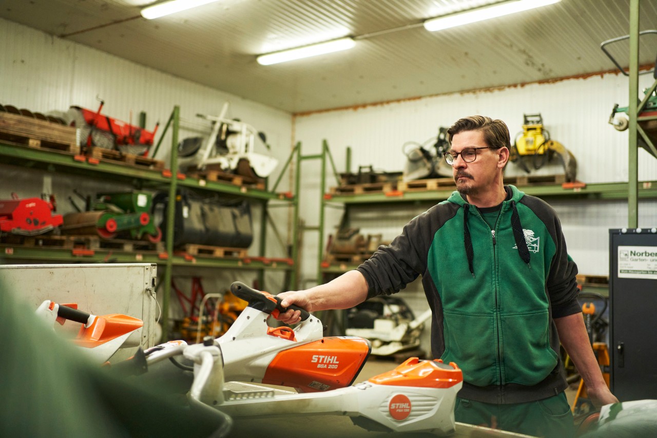 Мъж в сервиз стои зад тезгях с акумулаторни уреди STIHL, той посяга към акумулаторната духалка за листа STIHL BGA 200
