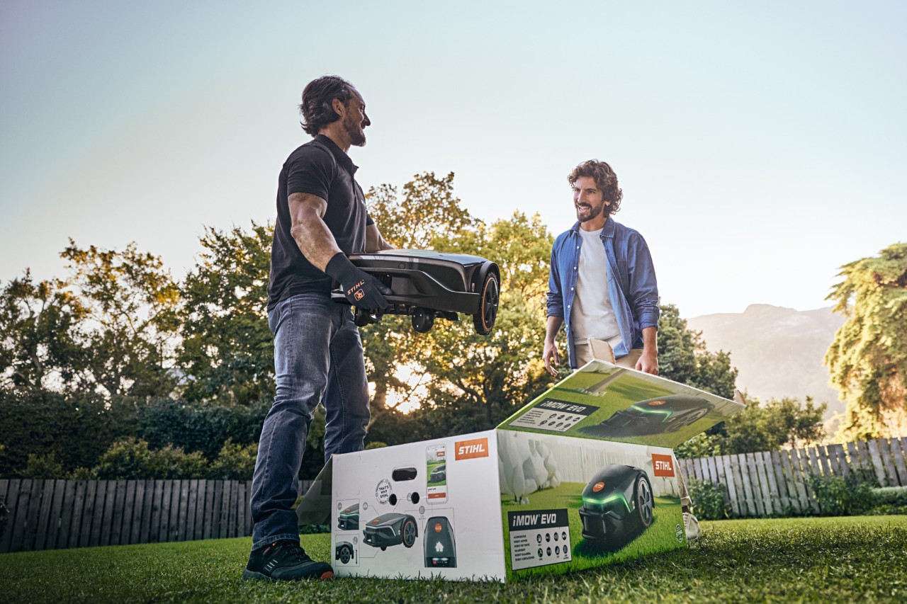 Специализиран търговец на STIHL стои с iMOW® EVO в ръка и iMOW® кашон пред него в градината на клиент и иска да помогне при инсталирането