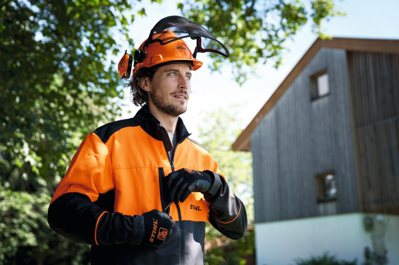 Мъж с оранжеви-черни предпазни средства STIHL, включително каска и ръкавици, гледа уверено от снимката, на заден план има къща и дървета 