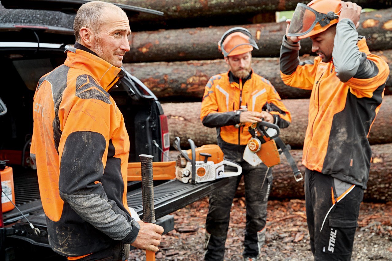 Трима мъже в защитно облекло STIHL пред купчина дърва се подготвят за работа с верижен трион STIHL